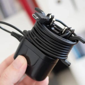 May include: A black power adapter with a coiled black cable and a black plastic clip. The cable is wrapped around the adapter.