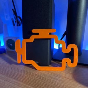 Pode incluir: Símbolo de luz do motor do carro de plástico laranja em uma superfície de madeira. O símbolo é uma representação estilizada de um motor de carro com uma luz de controle do motor.