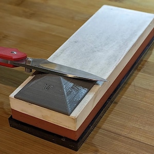 May include: A close-up of a two-sided sharpening stone with a gray pyramid-shaped honing guide. A red and silver pocket knife is resting on the stone.