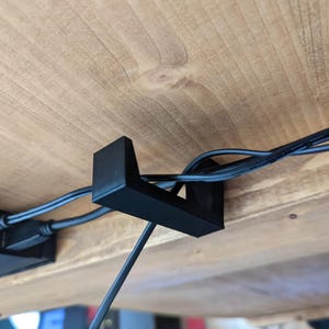 Puede incluir: Un clip de gestión de cables de plástico negro fijado a la parte inferior de un escritorio de madera, sujetando varios cables negros en su lugar.