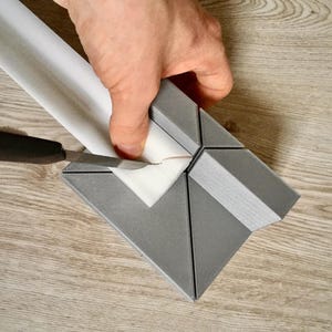 May include: A gray plastic tool for cutting miter joints in trim. A person is using the tool to cut a piece of white trim.