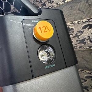 Op de afbeelding: Een close-up van een zwart-grijs powerstation met een oranje 12V-knop en een LED-lampje. Het powerstation heeft een handvat en een zilveren basis. De tekst "CIGAR" is zichtbaar boven de knop.