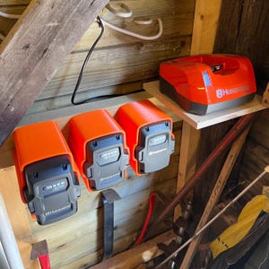 Może przedstawiać: Trzy pomarańczowe i czarne akumulatory Husqvarna są zamontowane na drewnianej ścianie. Czerwona ładowarka do akumulatorów Husqvarna znajduje się na półce nad akumulatorami.