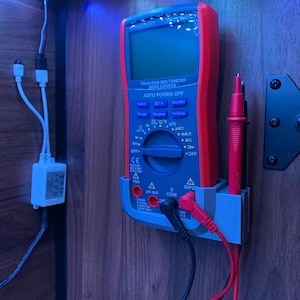 Op de afbeelding: Een rode en grijze digitale multimeter met een zwarte en rode sonde eraan bevestigd. De multimeter is gemonteerd aan een muur met een grijze houder. De tekst op de multimeter luidt "TRUE-RMS MULTIMETER 6000 COUNTS AUTO POWER OFF".