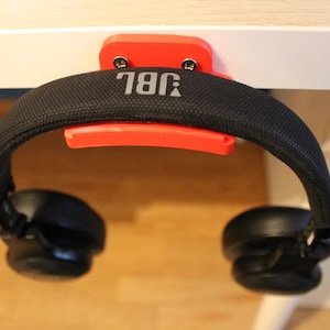 Puede incluir: Un soporte para auriculares de plástico rojo con la palabra "JBL" impresa en él. El soporte está fijado a un escritorio blanco y unos auriculares negros están colgados en él.