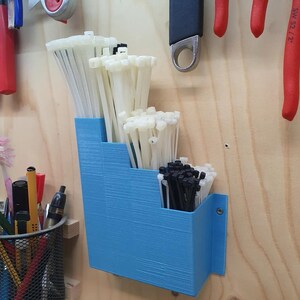 May include: A blue plastic wall-mounted organizer with three compartments for storing cable ties. The organizer is filled with white and black cable ties.