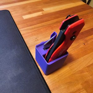 May include: A purple plastic holder with a red and black utility knife inside. The holder is sitting on a wooden surface.