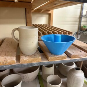以下が含まれることがあります： 木製の棚に展示された、マグカップ、ボウル、花瓶など、さまざまな陶器のコレクション。青いプラスチック製の型もあります。陶器はさまざまな段階で完成しており、陶芸の技術を示しています。