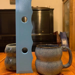 Puede incluir: Dos tazas de cerámica azul con un esmalte texturizado y moteado. Una taza tiene una forma redondeada y bulbosa, mientras que la otra tiene una forma de taza más tradicional. También se ve una herramienta de plástico azul claro con tres agujeros en la imagen.