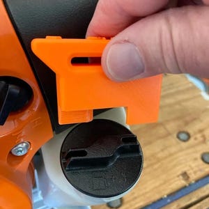 Puede incluir: Una herramienta de liberación de la tapa del tanque de combustible de plástico naranja para un soplador de hojas a gasolina. La herramienta está siendo sostenida por una mano y se está utilizando para liberar la tapa del tanque de combustible.