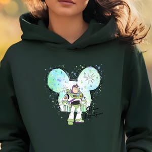 K&ouml;nnte beinhalten: Gr&uuml;ner Hoodie mit einer bunten Mickey-Maus-Silhouette, die Buzz Lightyear aus Toy Story zeigt. Der Text "Buzz Lightyear" ist in Kursivschrift unter dem Bild geschrieben.