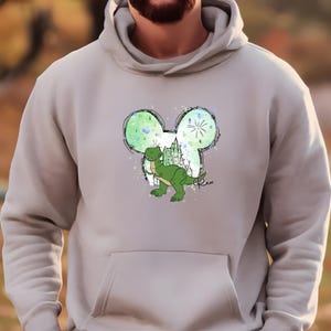 K&ouml;nnte beinhalten: Ein hellgrauer Hoodie mit einem gr&uuml;nen Dinosaurier in einer Mickey-Maus-Kopf-Silhouette. Der Dinosaurier ist gr&uuml;n und hat einen wei&szlig;en Hintergrund mit einem Schloss und Feuerwerk. Der Text "Rex" steht unter der Silhouette.