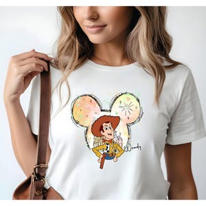K&ouml;nnte beinhalten: Wei&szlig;es T-Shirt mit einem farbenfrohen Aquarell-Mickey-Mouse-Kopf-Design mit Woody aus Toy Story. Das Design beinhaltet ein Feuerwerk und das Wort "Woody" in Schreibschrift.