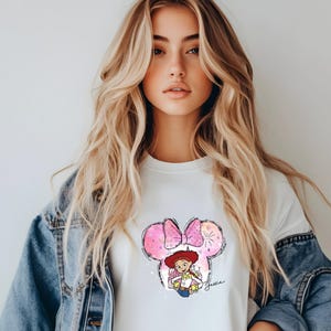 K&ouml;nnte beinhalten: Wei&szlig;es T-Shirt mit einem rosa und wei&szlig;en Aquarelldesign eines Comic-Charakters mit rotem Hut und Schleife. Der Charakter h&auml;lt ein Spielzeug und der Text "Jessie" steht unter dem Design.