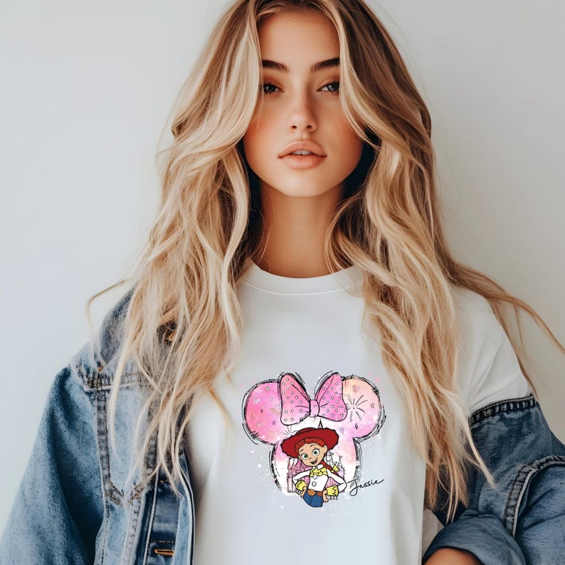 K&ouml;nnte beinhalten: Wei&szlig;es T-Shirt mit einem rosa und wei&szlig;en Aquarelldesign eines Comic-Charakters mit rotem Hut und Schleife. Der Charakter h&auml;lt ein Spielzeug und der Text "Jessie" steht unter dem Design.