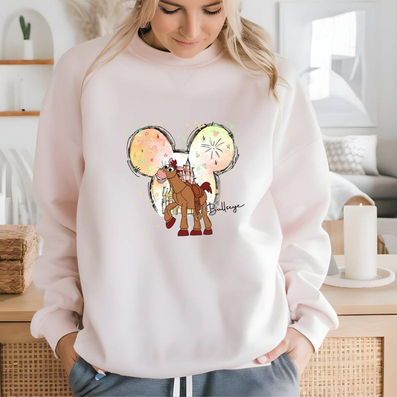 K&ouml;nnte beinhalten: Ein rosa Sweatshirt mit einer wei&szlig;en Mickey-Maus-Silhouette. Innerhalb der Silhouette befindet sich ein Cartoon-Bild eines braunen Pferdes mit Sattel und dem Text "Bullseye".
