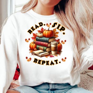 以下が含まれることがあります： 秋をテーマにしたグラフィックが施された白いスウェットシャツ。デザインはカボチャ、本、紅葉、コーヒーカップが特徴です。「READ. SIP. REPEAT.」のテキスト入り。読書好きや秋の愛好家に最適です。