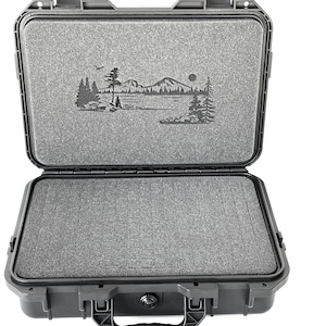 Può includere: Valigetta nera rigida con interno in schiuma e un design personalizzato inciso a laser che rappresenta una scena di montagna con alberi e un lago. La valigetta ha una maniglia e due chiusure.