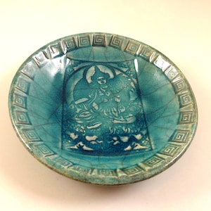 May include: A turquoise ceramic dish with a detailed embossed design of a seated figure. The dish has a crackle glaze and a geometric border. The central image is a detailed relief of a seated figure, likely a deity.