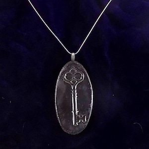 May include: A silver-colored key pendant on a thin silver chain. The pendant is an oval shape with a raised key design in the center. The key has a detailed, ornate head and a simple, rectangular keyhole. The background is a dark blue fabric.