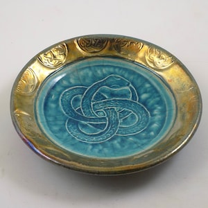 May include: Oval ceramic dish with a turquoise center featuring a white snake design. The dish has a gold-colored rim with embossed decorative elements. The dish is a decorative item.