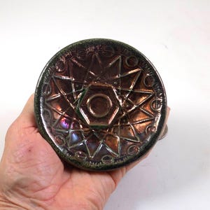 May include: A decorative, iridescent bottle stopper with a starburst design. The stopper features a central hexagon and is surrounded by a pattern of stars and circles. The color shifts from bronze to purple and green.