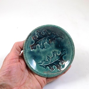 Può includere: Un piccolo piatto rotondo in ceramica turchese con due disegni di foglie di quercia in rilievo. Le foglie hanno dettagli marroni e il piatto ha un bordo strutturato. Il piatto è tenuto in mano su sfondo bianco.