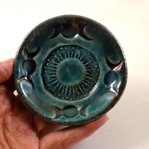 Op de afbeelding: Een kleine, turquoise keramische schaal met een verhoogd zonmotief in het midden en halve maanvormen langs de rand. De schaal heeft een glanzende afwerking en is ongeveer 10 cm in diameter.