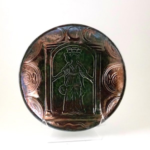 May include: A round, decorative plate with a central green image of a woman in a gown, framed by columns. The plate has a bronze-colored rim and embossed circular designs. The overall aesthetic is vintage and artistic.