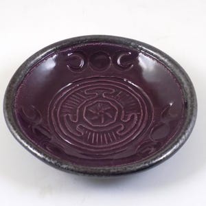May include: A small, purple ceramic dish with a silver rim. The dish features a design of a circle with a star in the centre and three crescent moons around the outside. The words "OOC" are written above the circle.