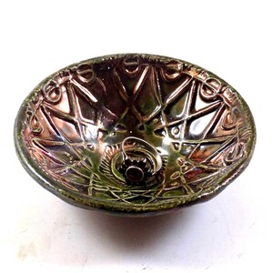 May include: A handcrafted ceramic bowl with an oval shape and a glossy, iridescent finish. The bowl features an intricate, raised geometric pattern in shades of green, brown, and bronze. A small, decorative ring sits in the center.