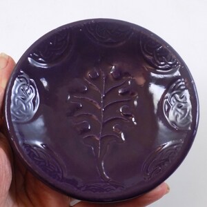 Può includere: Un piccolo piatto rotondo in ceramica di colore viola con un disegno in rilievo di una foglia di quercia al centro. Il piatto ha una finitura lucida ed è decorato con un motivo ripetitivo di foglie stilizzate attorno al bordo.
