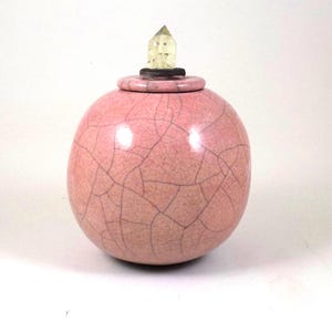 Può includere: Un vaso di ceramica rosa con finitura craquelé. Il vaso rotondo è sormontato da un coperchio e una punta di cristallo trasparente. Oggetto decorativo con sottili crepe.