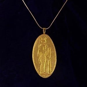 May include: Gold-colored pendant necklace featuring an oval pendant with an embossed Egyptian figure holding a staff. The pendant hangs from a thin, gold-colored chain. The background is a dark blue fabric.
