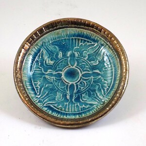 May include: A small, teal blue ceramic dish with a gold rim. The dish has a raised, embossed design of a stylized flower with a sunburst pattern.