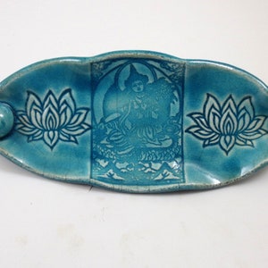 May include: A turquoise ceramic incense holder with a lotus flower design on each end and a central image of a seated figure. The holder has a small, round incense burner on one end.