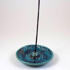 May include: A teal ceramic incense holder with a floral design. The holder has a small, raised center for the incense stick and a dark brown incense stick is inserted. The holder is round and has a glossy finish.