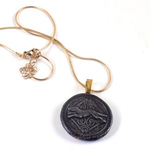May include: A gold-toned snake chain necklace featuring a circular pendant. The pendant has a dark, textured background with a raised silver-toned relief of a running greyhound or similar canine figure centered within a geometric border pattern.