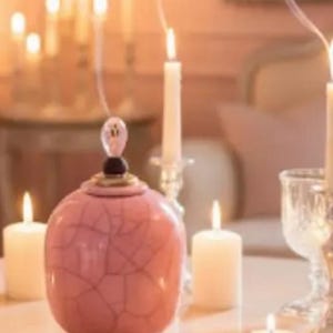 May include: A pink decorative jar with a cracked pattern, topped with a decorative finial, sits on a table. Lit candles of varying sizes and shapes surround the jar, creating a warm ambiance. Smoke rises from some of the candles.