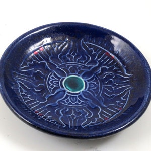 May include: A round, dark blue ceramic dish with a detailed white floral design. The center features a teal-colored gem. The dish has a glossy finish and is likely handmade. The dish is a decorative item.