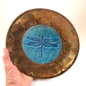 May include: A handmade ceramic plate with a blue dragonfly design in the center. The dragonfly is set against a cracked turquoise background. The plate has a bronze-colored border with spiral designs. The plate is round.