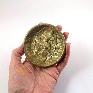Può includere: Un piccolo piatto rotondo e decorativo con un motivo a foglie in rilievo. Il piatto è dipinto in tonalità oro e verde, con una superficie strutturata. Il piatto è tenuto in mano su sfondo bianco.