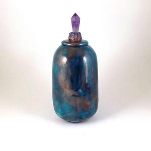 May include: A handcrafted, ceramic bottle with a teal and bronze metallic glaze. The bottle is topped with a decorative lid and a purple crystal. The bottle has a rounded, oval shape.