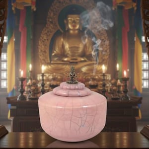 Puede incluir: Una urna de cerámica rosa con esmalte craquelado y tapa escalonada, coronada por una pequeña estatua. La urna está frente a una estatua dorada de Buda y velas, lo que sugiere un propósito espiritual o decorativo.