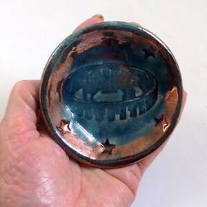 May include: A small, handmade ceramic bowl with a teal interior and a bronze-colored rim. The bowl features a raised design in the center and star-shaped cutouts around the edge. The bowl is held in a hand.