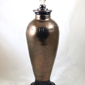 May include: A tall, bronze-coloured ceramic vase with a black base and a crystal diamond-shaped stopper. The vase has a textured surface and a decorative band around the middle.