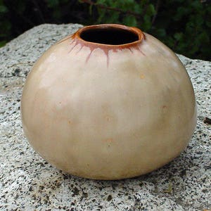 May include: A small, round, beige ceramic vase with a dark brown interior and rim. The vase has a smooth surface and sits on a gray, textured rock. The rim has a brown, drip-like design. The vase is a decorative item.
