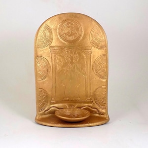 May include: A gold-colored, arched wall hanging with a relief design. The design includes a central figure within an architectural frame, surrounded by circular patterns. A small, round basin is at the base. The piece is likely a decorative item.