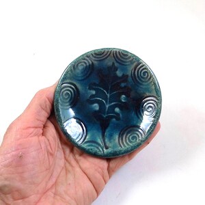 Può includere: Un piccolo piatto rotondo in ceramica con smalto turchese. Presenta un motivo a foglia blu scuro al centro e motivi a spirale sul bordo. Il piatto misura circa 7,6 cm di diametro.