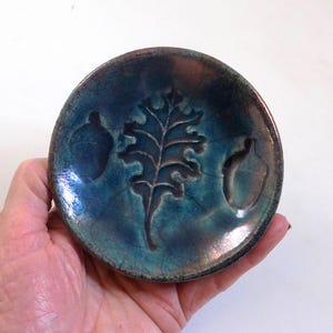 Può includere: Un piccolo piatto rotondo in ceramica con smalto verde acqua e marrone. Il piatto presenta una foglia di quercia in rilievo e due ghiande. Il piatto è tenuto in mano da una persona.